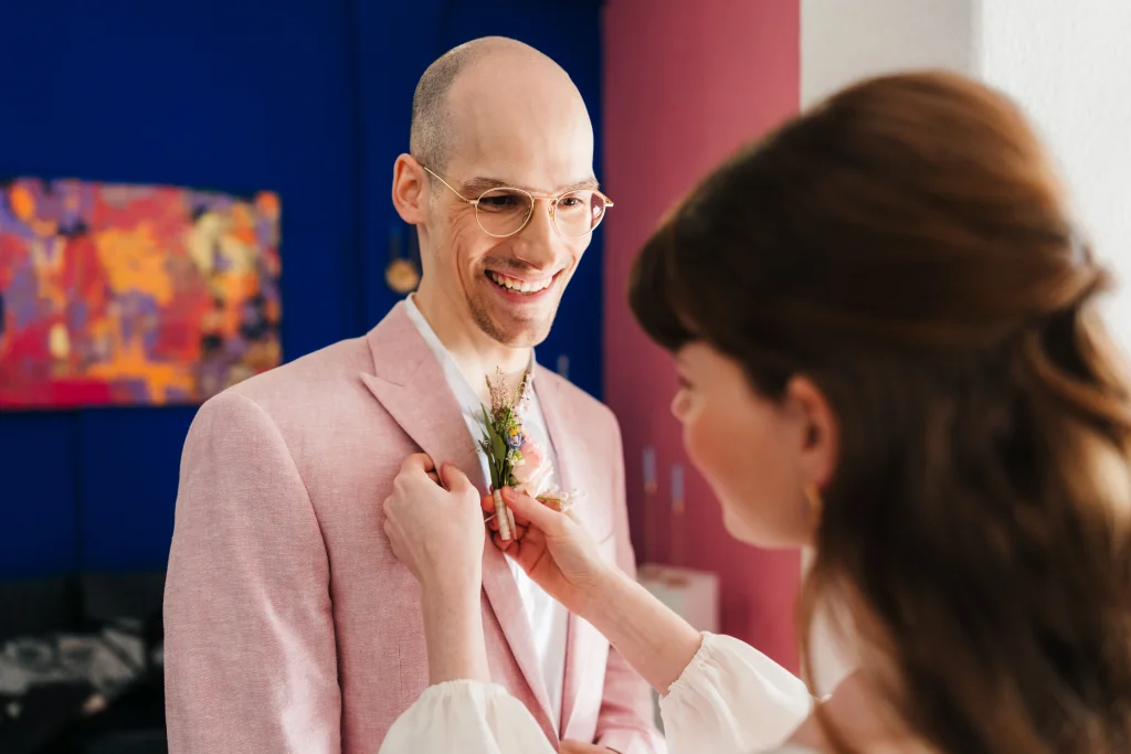 Bride pins a boutonniere on the lapel of her groom’s pink suit