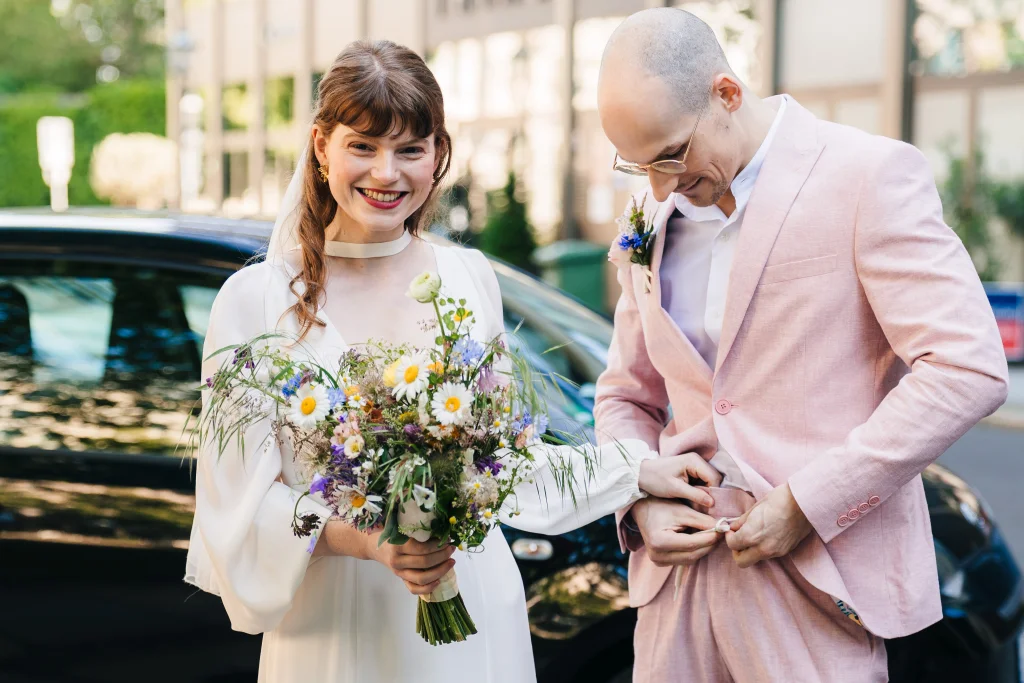 Bride in a white dress holds a colorful bouquet of flowers while her groom in a pink suit adjusts his belt.