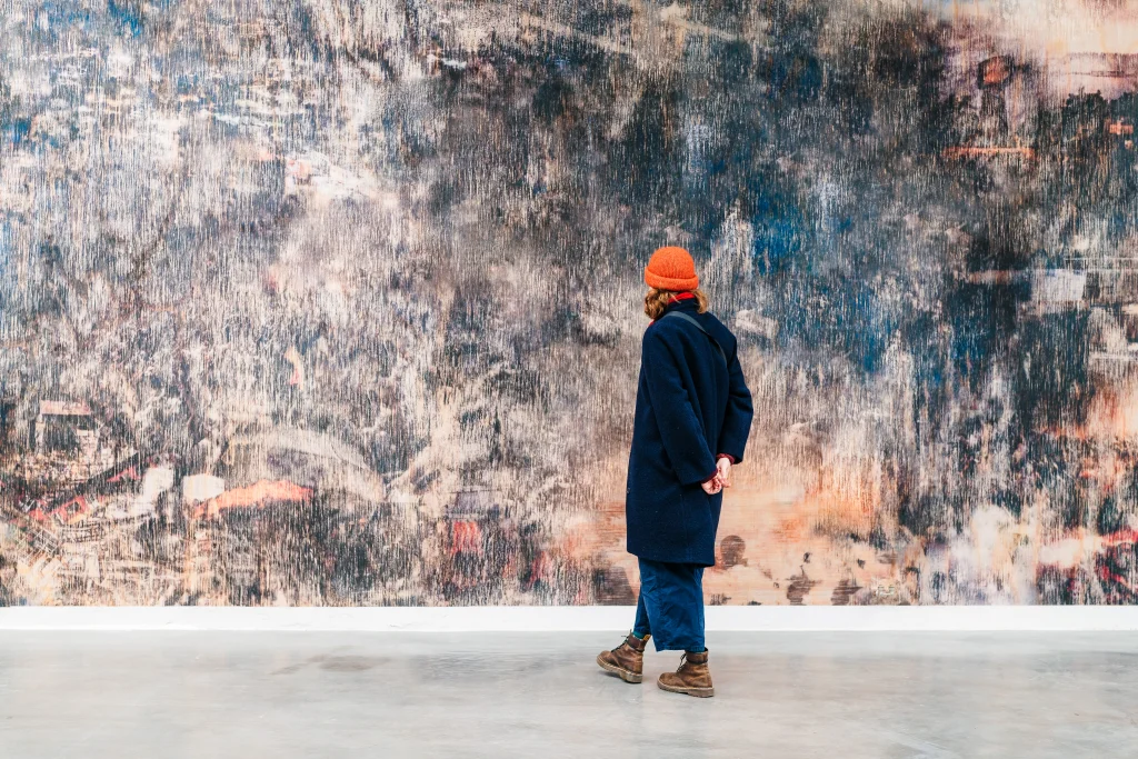 Person wearing a dark coat and orange beanie standing in front of a large abstract textured wall painting