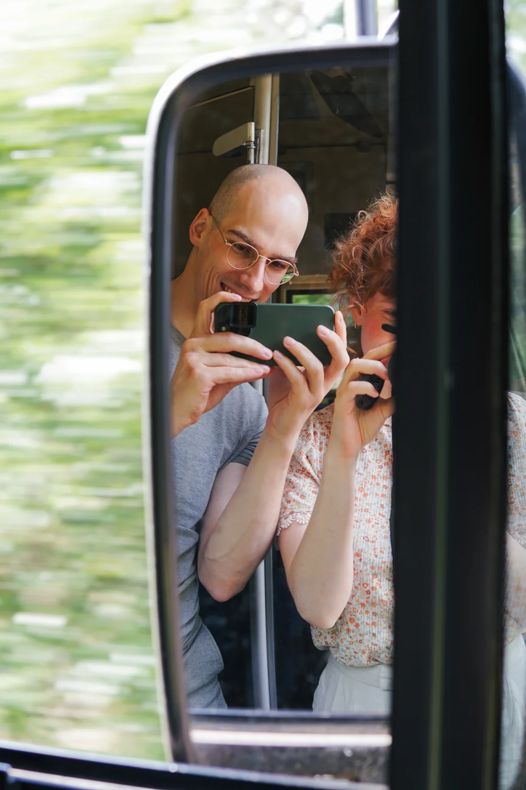 Reflection of two people smiling and taking photos in a vehicle’s side mirror