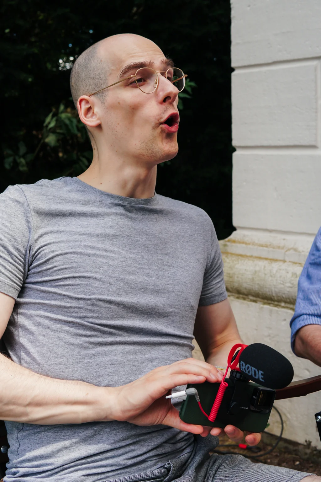 Person in a gray shirt speaking while holding a small camera setup with an attached microphone